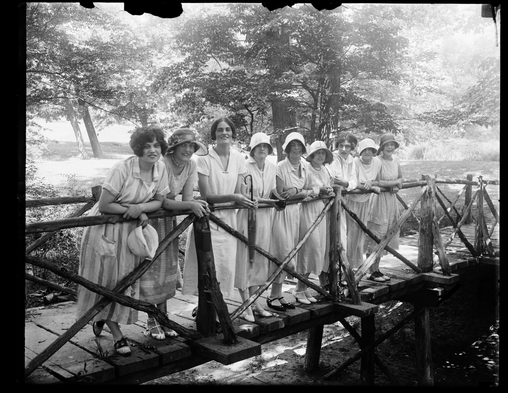 Nine young women in stylish 1920s white dresses pose on a rustic bridge in a park