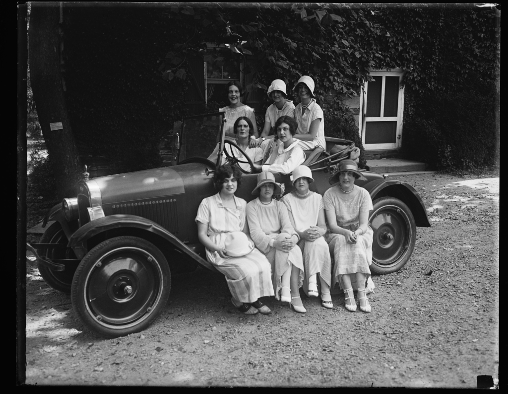 Young women in 1920s dress pose in a car in front of Peirce Mill